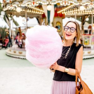 Young woman standing with pink cotton candy outdoors in front of the carrousel at the amusement park